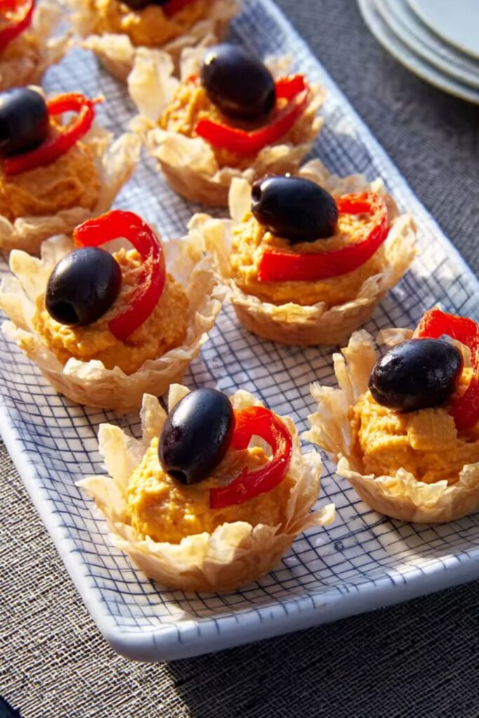 A tray of spooky Halloween snacks, specifically Slithering Hummus Bites, featuring bite-sized phyllo cups filled with hummus and topped with a black olive.