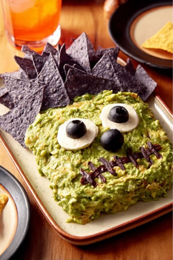 Guacamole shaped into a monster face with tortilla chips for hair, decorated with sour cream, black beans, and olives as creepy Halloween snacks.