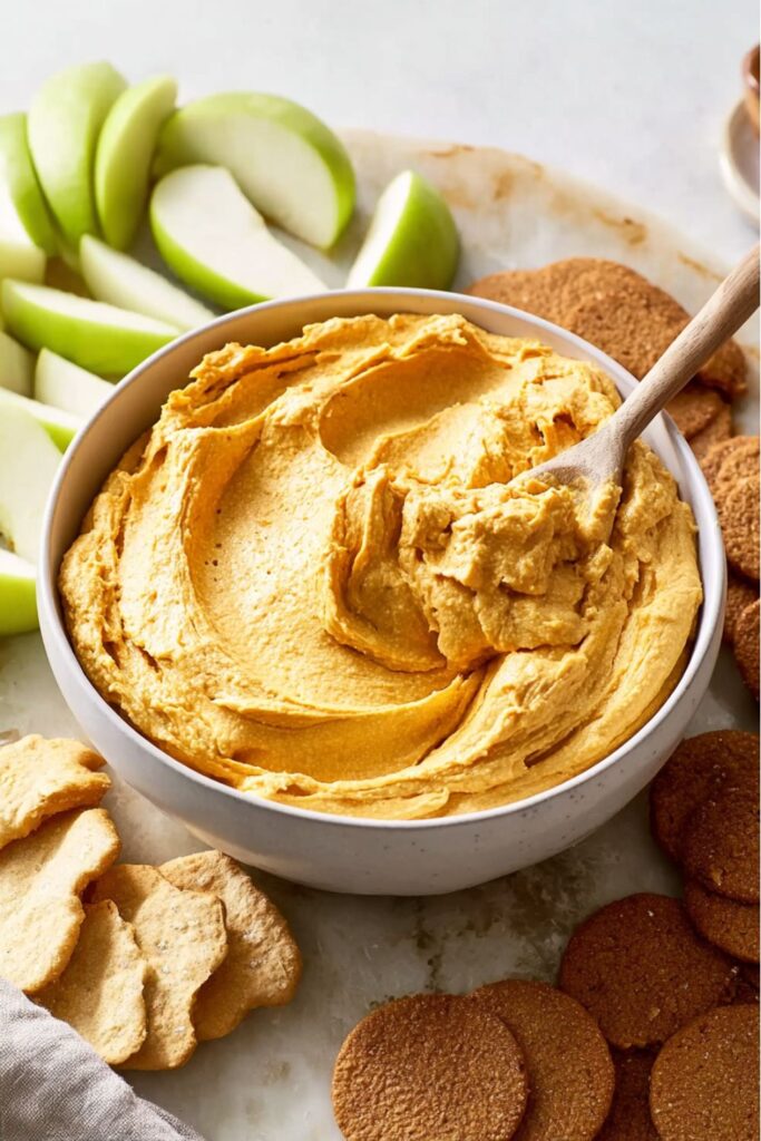 A creamy pumpkin dip served in a glass bowl on a marble countertop, topped with a dusting of pumpkin spice, surrounded by cookies and crackers as Halloween finger foods.
