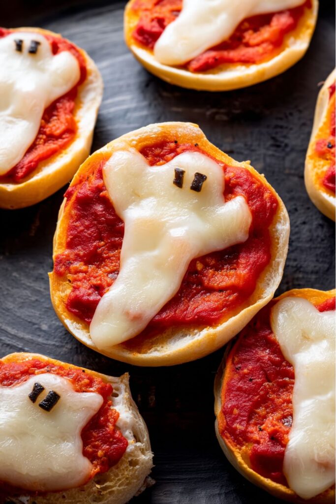 Mini bagels topped with marinara, melted mozzarella shaped like ghosts, and tiny olive eyes, styled as spooky Halloween finger foods.