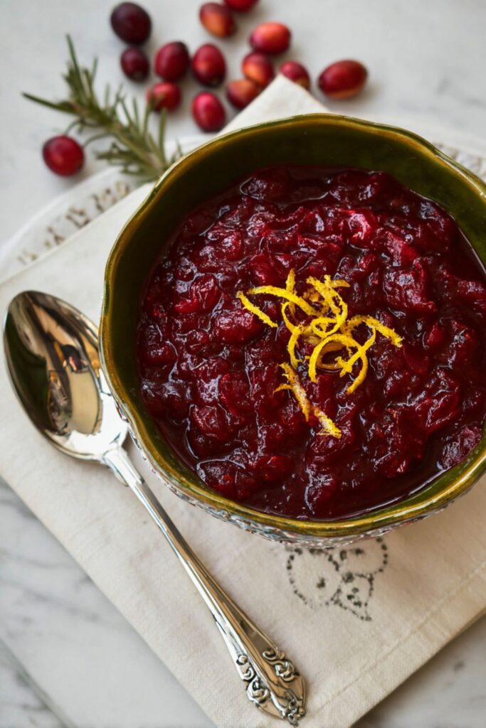 A bowl of homemade cranberry sauce from the best Ina Garten recipes, showing vibrant red cranberries with chunks of apple, garnished with orange and lemon zest, set on a marble countertop.
