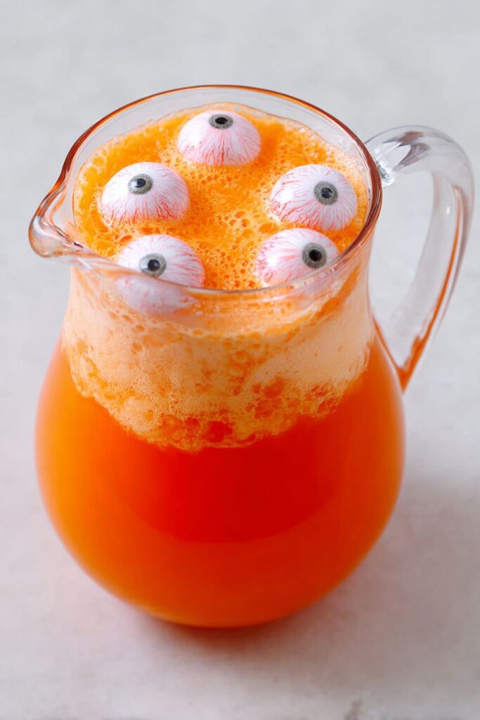A bright orange punch bowl of Halloween batch cocktails made with soda, pineapple juice, and scoops of orange sherbet, decorated with floating plastic eyeballs for a spooky effect.