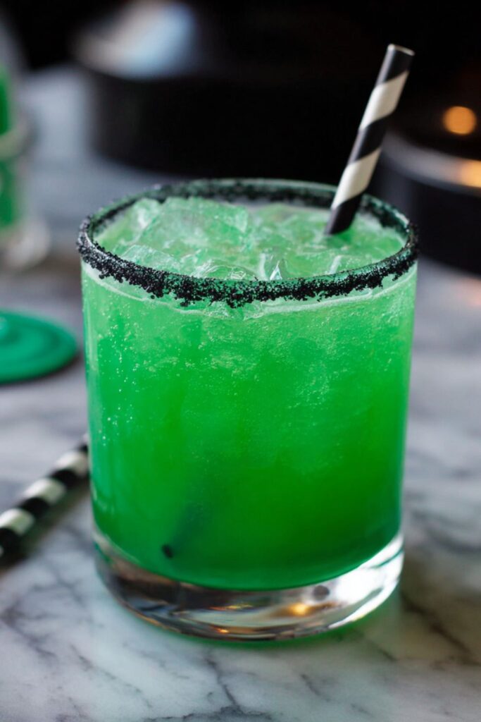 A glowing green Halloween cocktail in a rocks glass rimmed with black salt, filled with ice and garnished with a striped straw, perfect for spooky Halloween themed drinks.