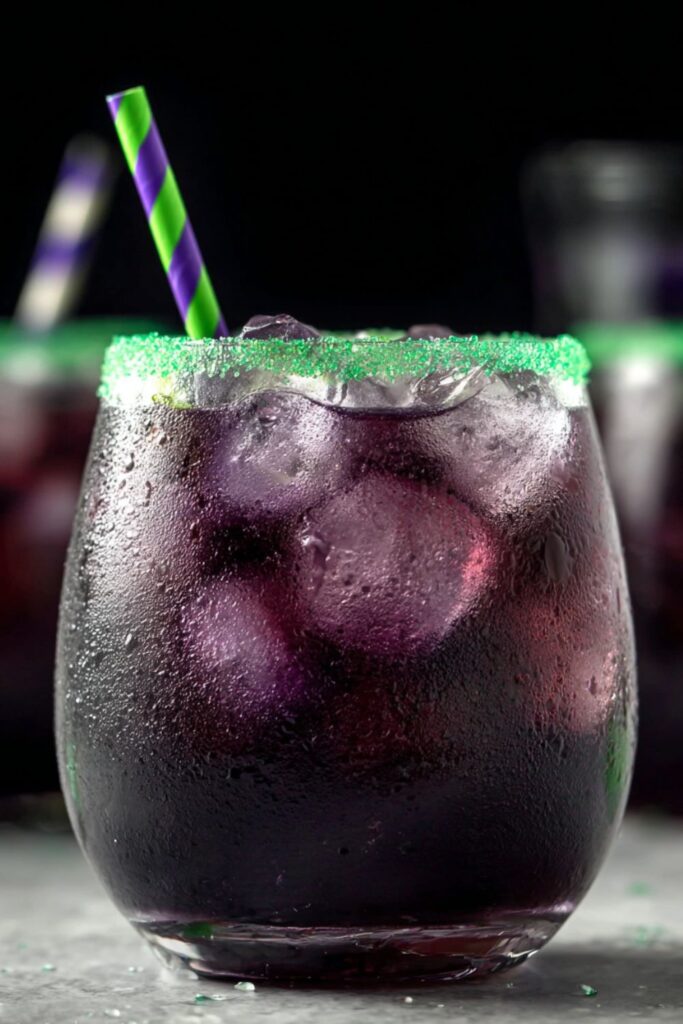 A tumbler filled with layered blue and red Halloween cocktail over ice, topped with soda and star sprinkles, rimmed with green sugar for a spooky Halloween themed drink.