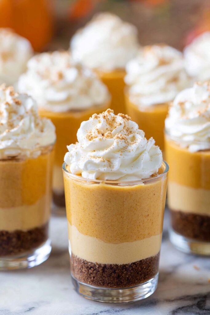 Mini pumpkin pie cheesecake shooters layered with gingersnap crust, creamy pumpkin filling, and whipped cream, served in small glasses as simple Thanksgiving desserts.