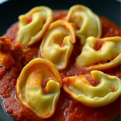 a modern dark plate of ear-shaped beef ravioli golden pasta pouches with crimped rims, nestled in glossy red marinara, steam rising, creamy ricotta-beef center, tender silky texture.