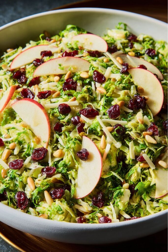 Shaved Brussels sprout salad with apple slices, shredded cheddar, dried cranberries, and pine nuts tossed in a light apple cider vinaigrette, served as a refreshing Thanksgiving vegetable side dish.