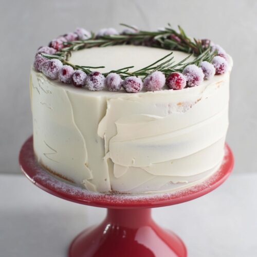 a white Christmas Minimalist cake with smooth, creamy frosting and a slightly rustic finish, topped with a neat circular wreath of sugared cranberries arranged along the edge and interwoven with fresh green rosemary sprigs, all set on a red pedestal against a soft gray background.