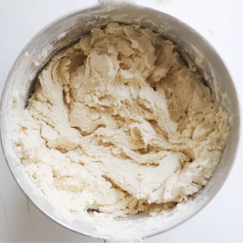 a large metal mixing bowl filled with a pale, sandy mixture that looks soft and fine to the touch. The blend has a uniform, crumbly texture with tiny buttery specks that catch the light, giving it a faint golden hue. The surface is slightly uneven, like smooth, sifted sand, showing the perfect combination of flour, sugar, baking powder, salt, and butter blended into a delicate, velvety base.