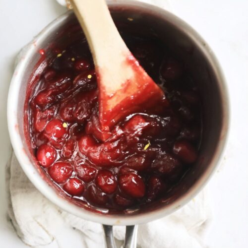 small stainless steel saucepan sitting on a light kitchen counter, filled with glossy ruby-red cranberry jam that glistens under soft natural light. The thick mixture has visible whole cranberries, some burst open, releasing deep crimson juices that swirl with a syrupy sheen. A wooden spoon rests inside the pot, coated with the vibrant jam, while faint steam rises, suggesting warmth.