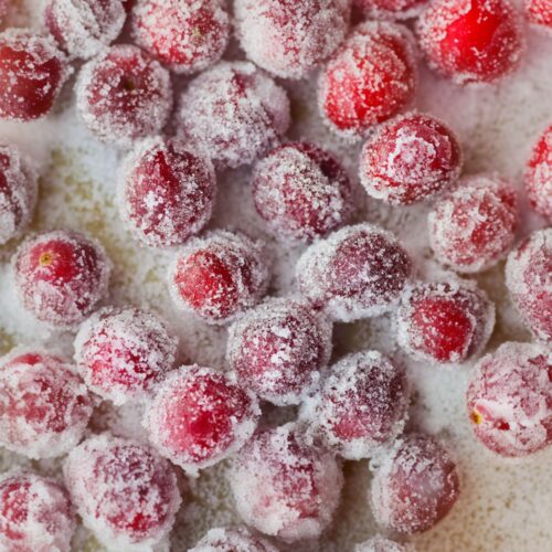 close-up view of glistening sugared cranberries, each one coated in a sparkling layer of fine sugar crystals. The deep red berries peek through the frosty coating, creating a mix of vibrant crimson and soft white tones. Their surfaces look crisp and slightly rough with the grainy sugar, while the light catches on the crystals, making them shimmer like tiny gems. The background is dusted with loose sugar, enhancing the wintry, festive look of these beautifully frosted cranberries.