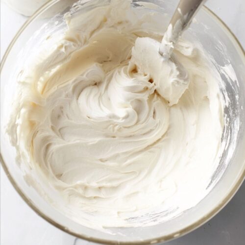 glass mixing bowl filled with thick, creamy white buttercream frosting, its texture smooth yet fluffy with soft peaks and gentle swirls from a spatula resting inside. The frosting has a glossy sheen, rich and velvety, showing subtle ridges where it’s been folded and spread.