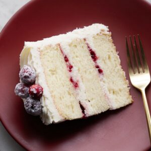 a single slice of white layer minimalist Christmas cake resting on a sleek red minimalist plate, its pale, tender crumb contrasting sharply with the dark surface. The cake has three soft layers filled with creamy white frosting and thin ribbons of crimson cranberry jam that peek through with a glossy sheen.