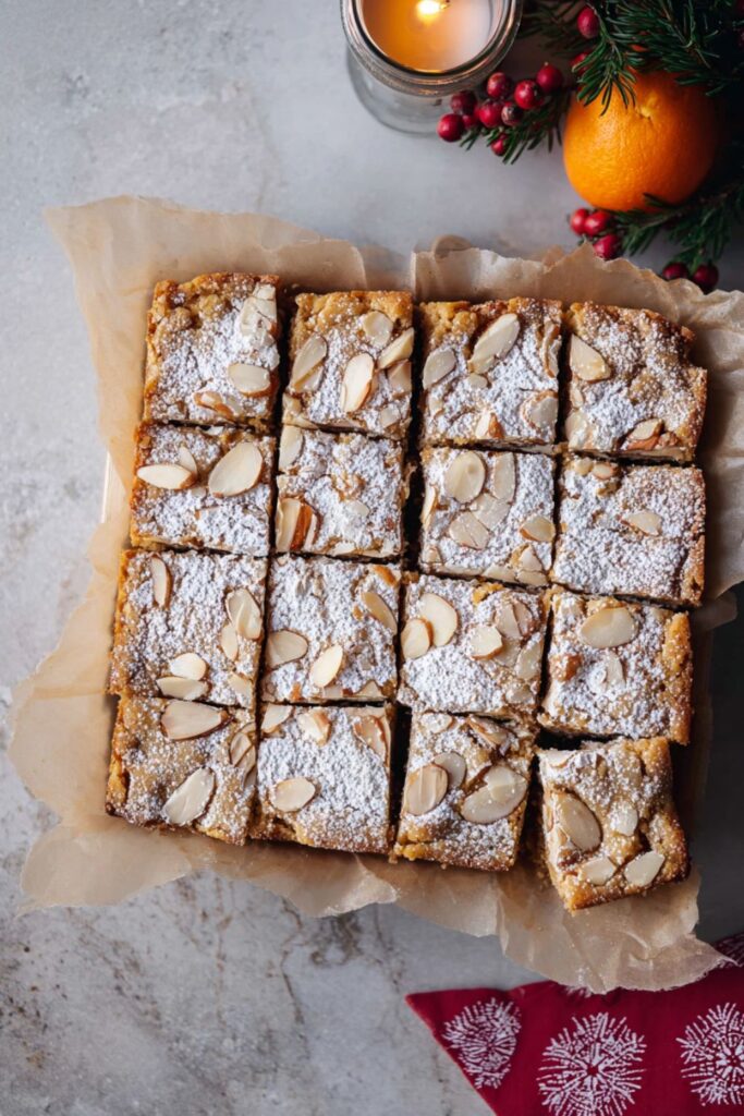 An overhead shot of several golden brown Christmas Bar And Square pieces, likely almond or frangipane bars, cut into neat squares. The Christmas Baking Recipe bars are topped with toasted slivered almonds and a dusting of white powdered sugar. They are arranged on parchment paper in a square pan or tray. The surrounding setting includes festive elements like a lit candle, an orange, red berries, and pine sprigs, suggesting a sweet Winter Holiday Dessert or Christmas Baked Good.