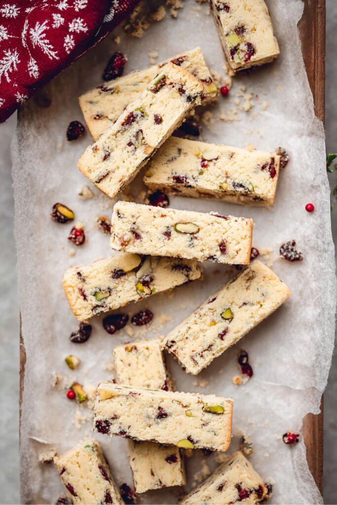 An overhead shot of several rectangular shortbread Christmas Cookie Bar pieces scattered across a piece of parchment paper on a wooden board. The Christmas Bar And Square pieces are light-colored and dense, studded throughout with bits of bright green pistachios and dark red dried cranberries, creating a festive color palette. Crumbs, loose nuts, and cranberries are visible around the bars, which are ready for a Holiday Baking display. A red snowflake-patterned napkin and small red berries frame the corner of the image, emphasizing the Winter Holiday Dessert theme.