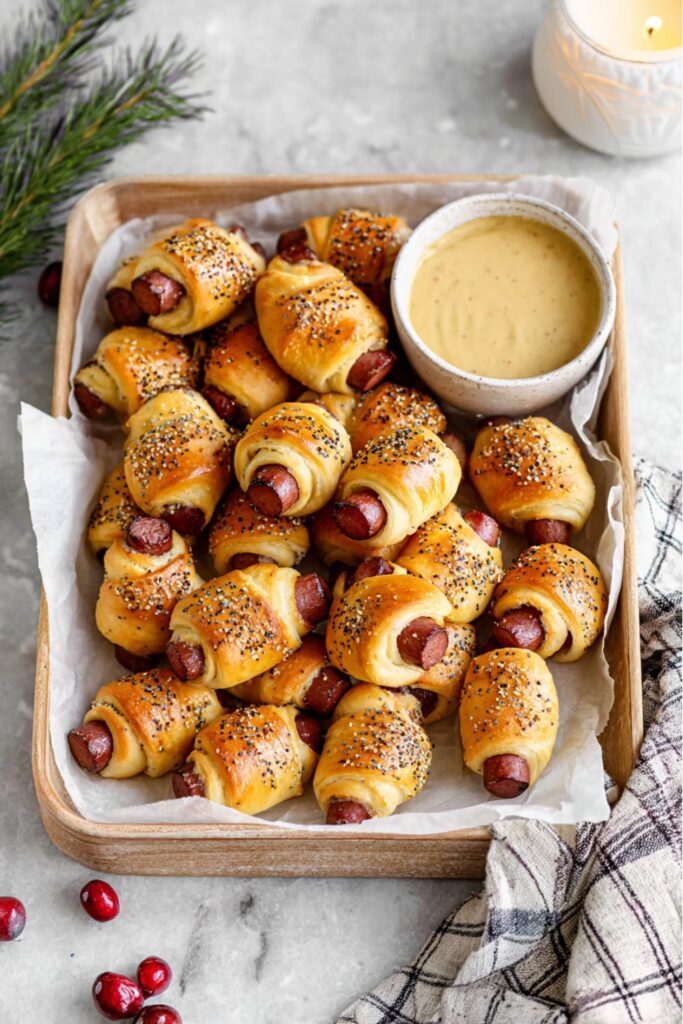 Golden-brown pigs in a blanket topped with everything bagel seasoning, served with a side of honey mustard dip on a marble countertop as festive Christmas party finger foods.