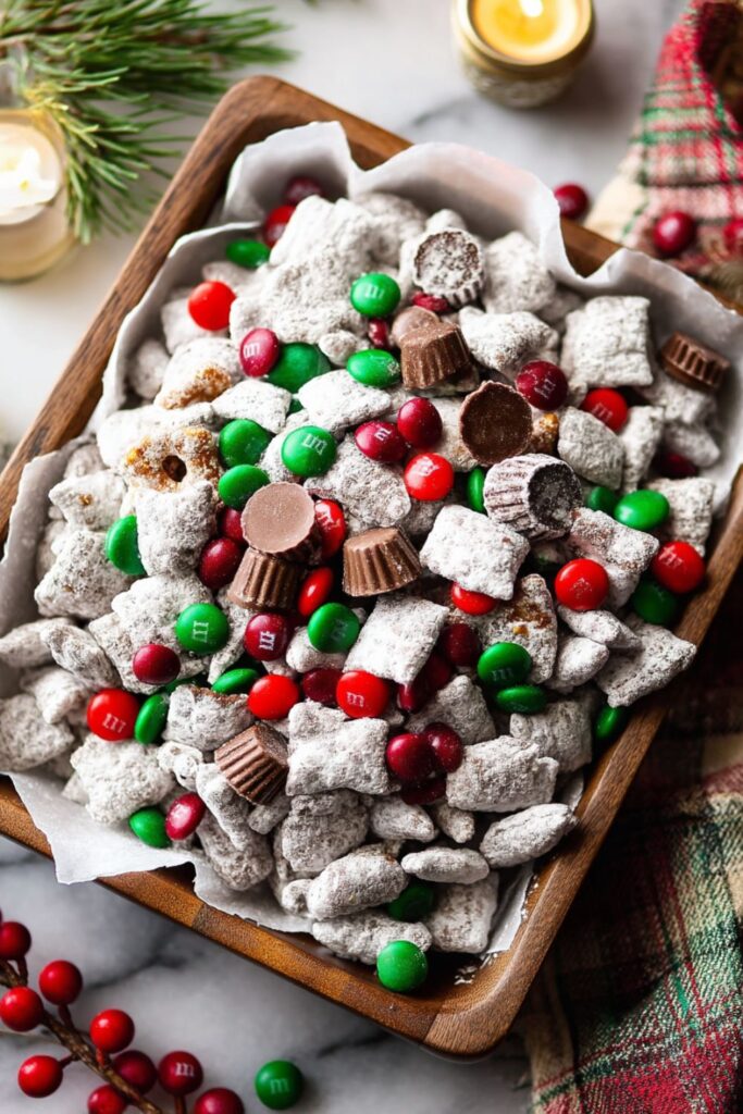A close-up of Christmas themed snacks served in a wooden bowl lined with white parchment paper. The snack is a batch of "Puppy Chow" or "Muddy Buddies" (Chex mix coated in chocolate, peanut butter, and powdered sugar) mixed with red and green M&M's and mini peanut butter cups. This sweet treat is a perfect Make Ahead Christmas Dessert or for Christmas Eve Dessert Ideas.
