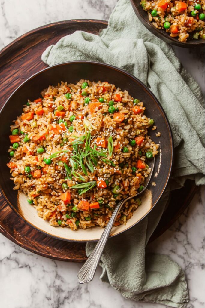 A dark bowl of savory vegan fried rice, studded with bright orange carrots and green peas, topped with sliced scallions and sesame seeds, illustrating an Easy Vegan dinner.