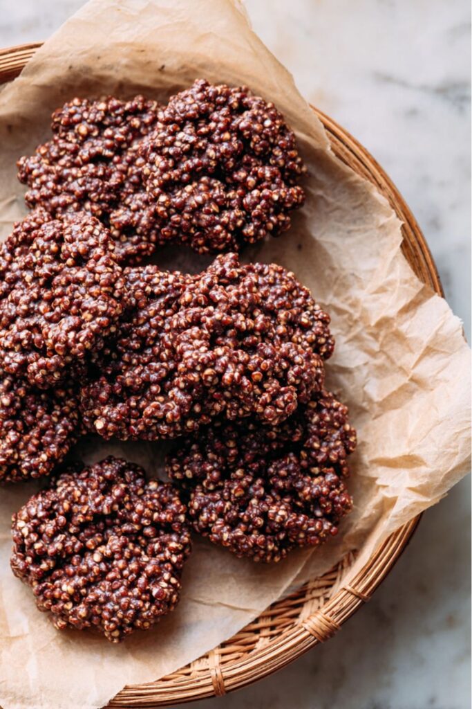 A close-up of several dark, chocolate-brown puffed quinoa clusters, shaped like cookies, resting on a sheet of crumpled parchment paper inside a woven basket, representing a delicious healthy no bake cookie.