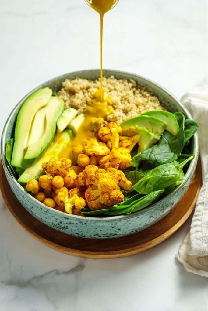 A close-up view of a vibrant Quinoa Salad Meal Prep Healthy Lunches bowl filled with fluffy quinoa, fresh spinach, sliced avocado, chickpeas, and roasted turmeric cauliflower florets, with a stream of yellow turmeric dressing being poured over the top.