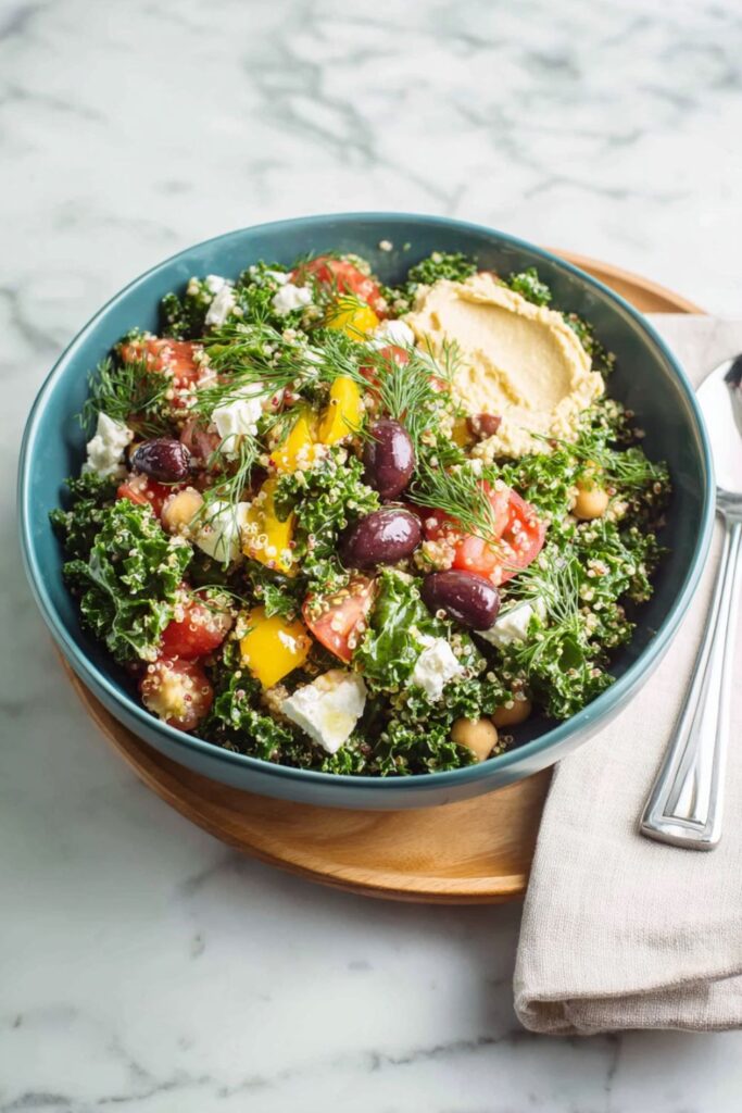A vibrant Quinoa Salad Lunch Prep in a blue bowl, featuring a mix of massaged kale, quinoa, chopped tomatoes, yellow bell peppers, Kalamata olives, crumbled feta cheese, and a dollop of hummus, garnished with fresh dill.