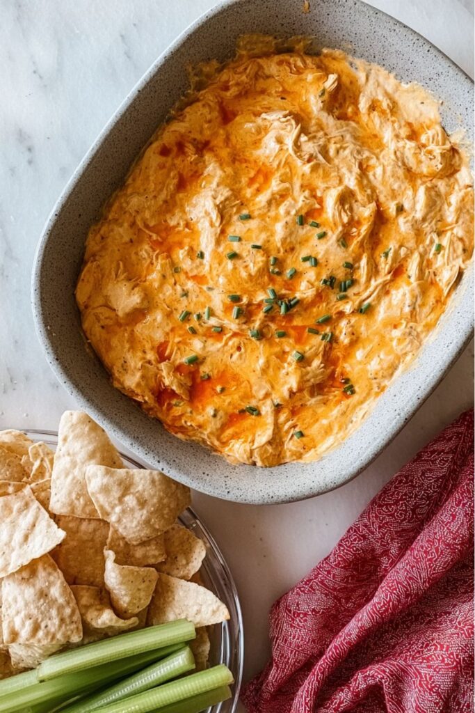 A bowl of creamy buffalo chicken dip topped with chives and served with tortilla chips and celery, perfect for anyone searching for healthy super bowl dips.