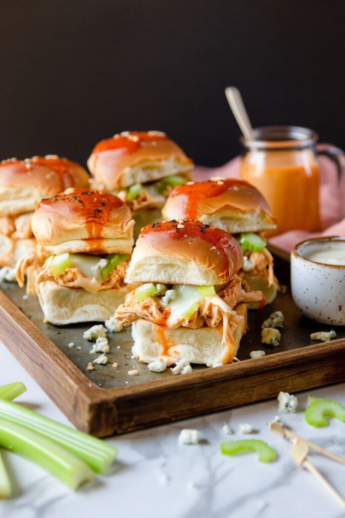 A tray of toasted pull-apart buffalo chicken sliders topped with blue cheese crumbles and sliced celery, ready for food spreads for parties.
