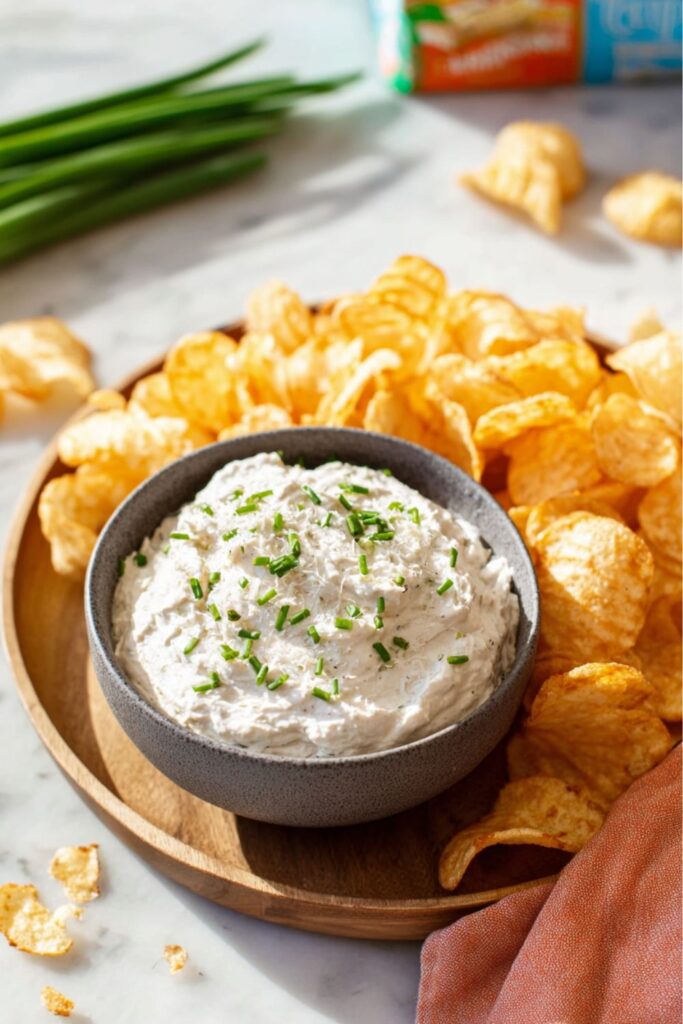 A bowl of creamy onion dip garnished with fresh chives and served on a wooden platter with potato chips, representing ideal dairy-free game day food.