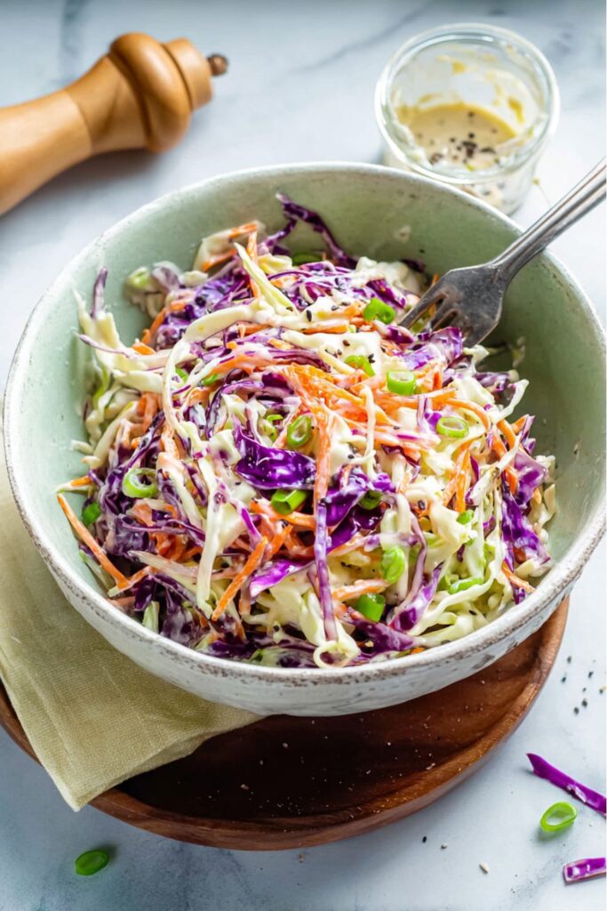 A bowl of vibrant green and purple shredded cabbage slaw tossed in a creamy dressing with sliced green onions, ideal for easy super bowl sides and food spreads for parties.