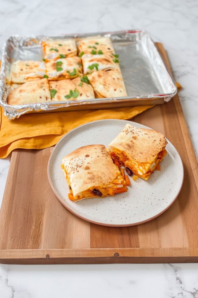 Two sliced portions of golden-brown sheet pan quesadillas filled with black beans and peppers on a plate, with the remaining tray of sliced quesadillas in the background for a football watch party.