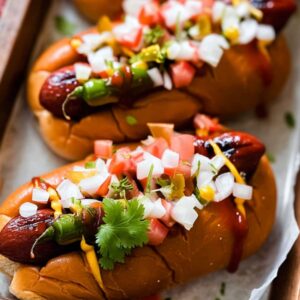 Loaded hot dogs with salsa, charred serranos, and cilantro on toasted buns in tray.