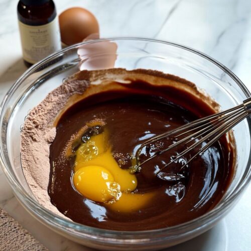 Chocolate cake batter in glass bowl with egg and whisk on marble countertop.