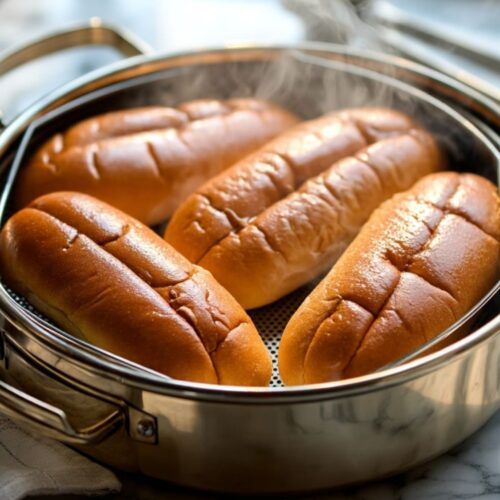 Steaming hot dog buns in metal steamer basket on marble countertop with soft natural light.