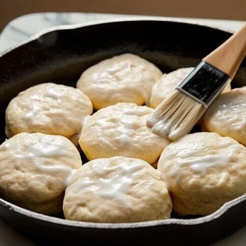 Pastry brush spreading buttermilk over biscuit dough rounds in cast iron skillet before baking.