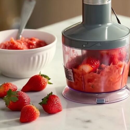 Portable food processor blending fresh strawberries on marble countertop with bowl of strawberry puree nearby.