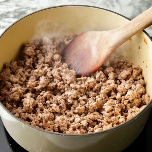 Crumbled ground beef and sausage browning in a Dutch oven on a marble countertop.