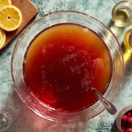 Punch recipe being mixed in a glass bowl with spoon, citrus slices, and ingredients nearby.