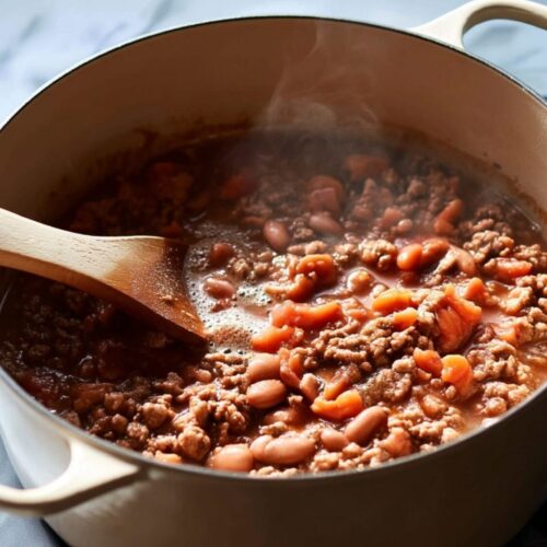 Beef, sausage, and beans simmering in a cream-colored Dutch oven on a marble countertop.