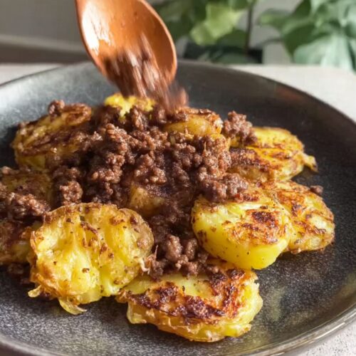 Cooked ground beef spooned onto crispy smashed potatoes on a dark artisan plate.