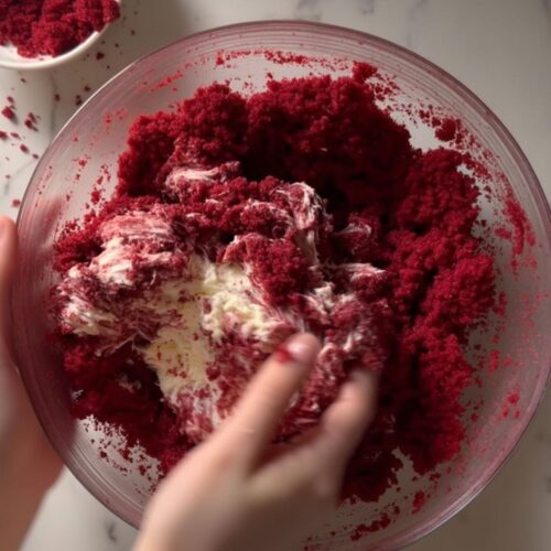 Hands mixing red velvet cake crumbs with cream cheese frosting in bowl for Cinco de Mayo desserts.