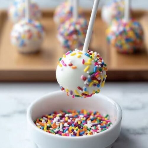 White chocolate cake pop dipping into rainbow sprinkles, with decorated cake pops on tray behind.