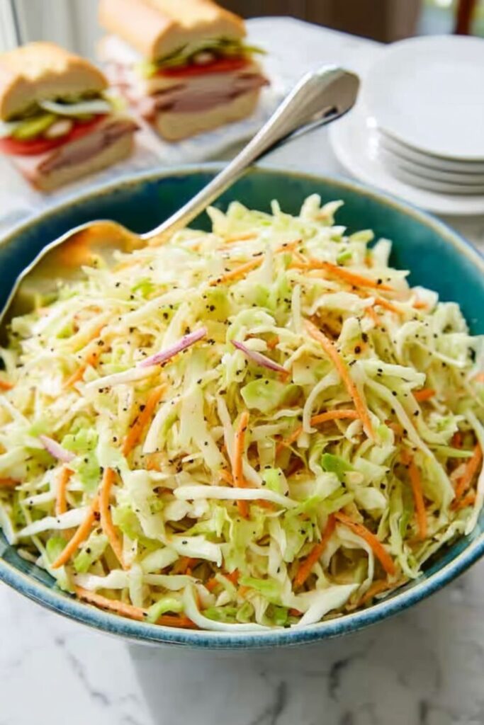 A blue bowl of vinegar-based green cabbage coleslaw with turkey subs behind it.