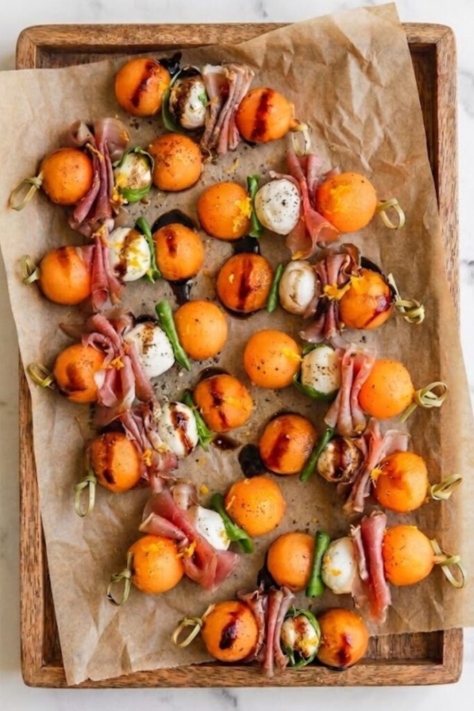 Cantaloupe melon, prosciutto, and mozzarella skewers drizzled with dark balsamic glaze on parchment paper.