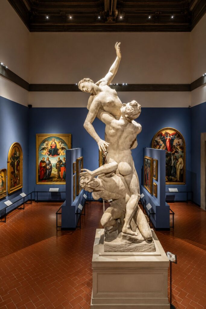 Marble sculpture in the Accademia Gallery with blue walls, Renaissance paintings, and spotlights