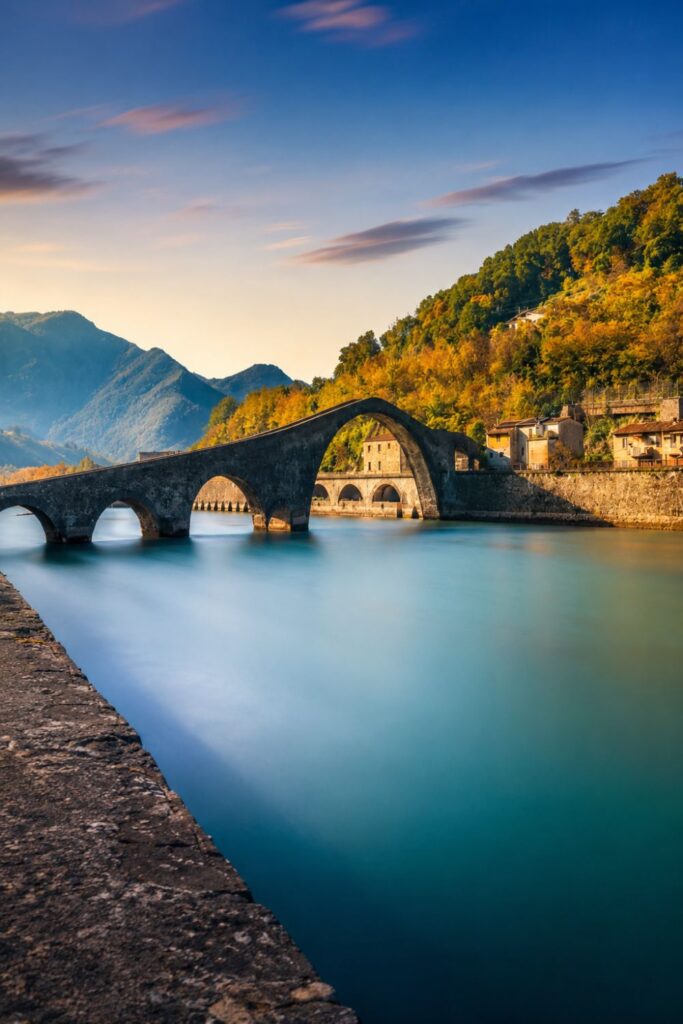 Stone arches of the Devil’s Bridge near Barga crossing calm water below wooded hills