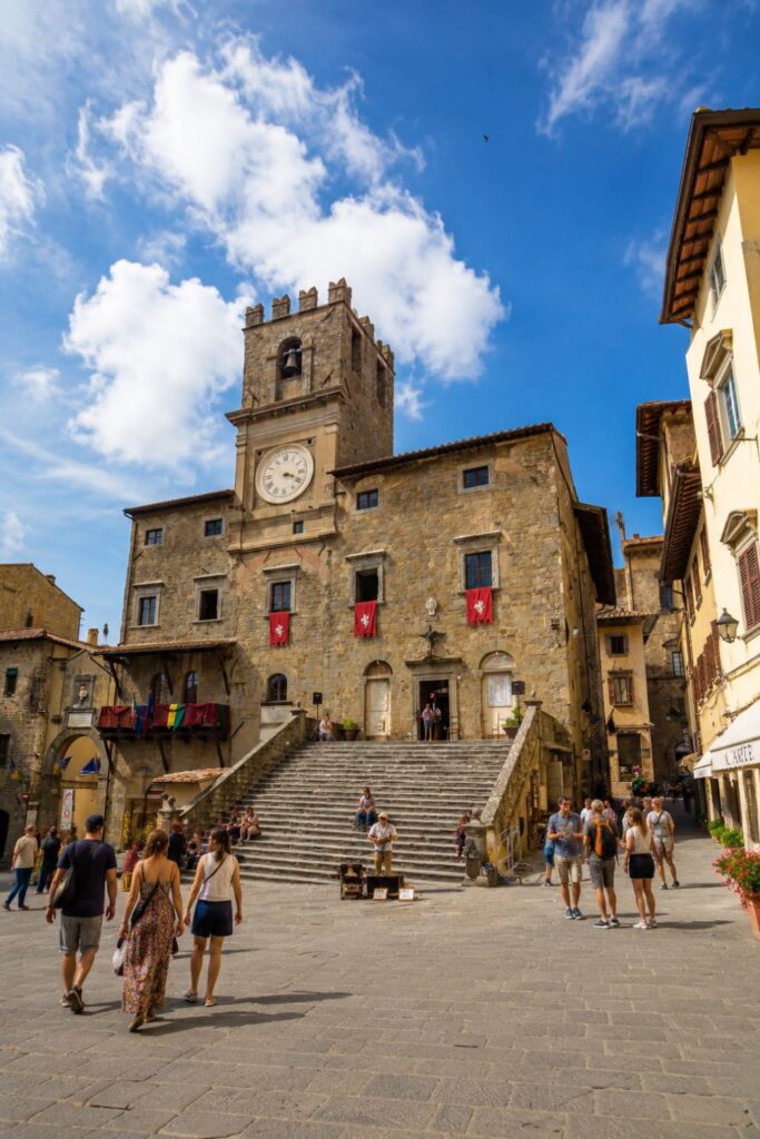 Cortona’s stone town hall, clock tower, broad steps, and sunny piazza with visitors
