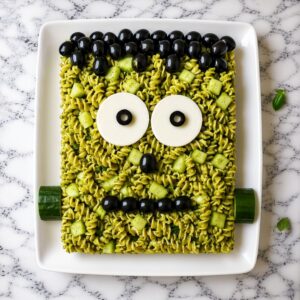 Frankenstein's monster face made from green pesto pasta, cheese, and olives on a plate.