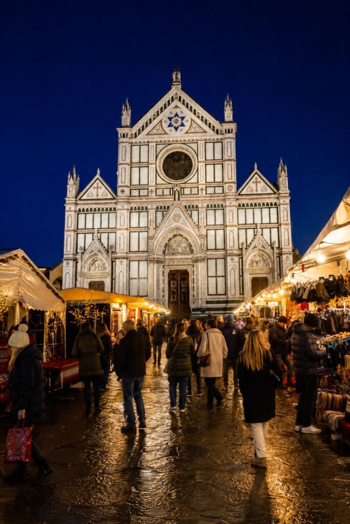 Christmas stalls glow beneath Santa Croce’s marble facade as winter shoppers cross the wet piazza.