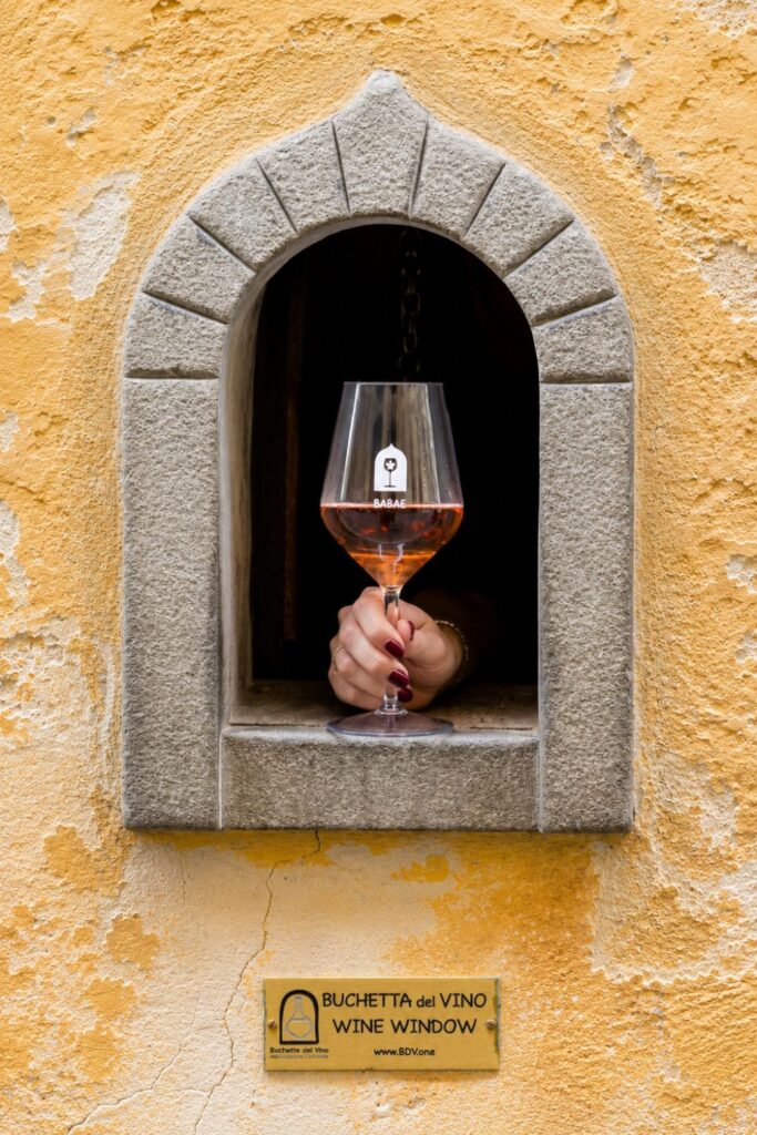 A hand presents rosé through a stone-framed wine window in a weathered ochre Florence wall.