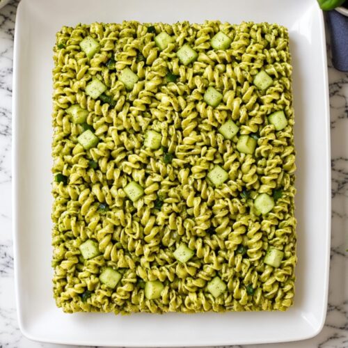 A rectangular block of green pesto rotini pasta mixed with cucumber pieces on a plate.